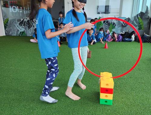 Two children guiding a blindfolded classmate through a trust exercise on the indoor turf.